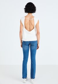 Hvid ærmeløs top med en dyb rygudskæring, parret med blå tætsiddende jeans og sølvfarvede sneakers, mod en ensfarvet baggrund.