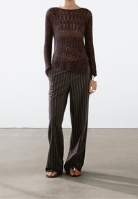 Haut marron à manches longues et côtelé avec un col bateau, associé à un pantalon large à rayures sombres. La tenue présente un design léger et texturé.