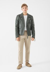 Grön läderbikerjacka med silverdragkedjor, en figursydd design och stora slag, kombinerad med ljus khakibyxor och beige skor.