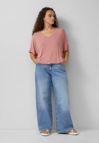 Roze geribbelde V-hals top met korte mouwen, gedragen met losse lichtblauwe jeans en witte sandalen, staand tegen een neutrale achtergrond.