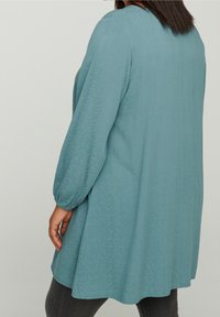 Tunique à manches longues en tissu bleu sarcelle doux avec une texture subtile, encolure arrondie et forme trapèze, portée sur un pantalon slim sombre.