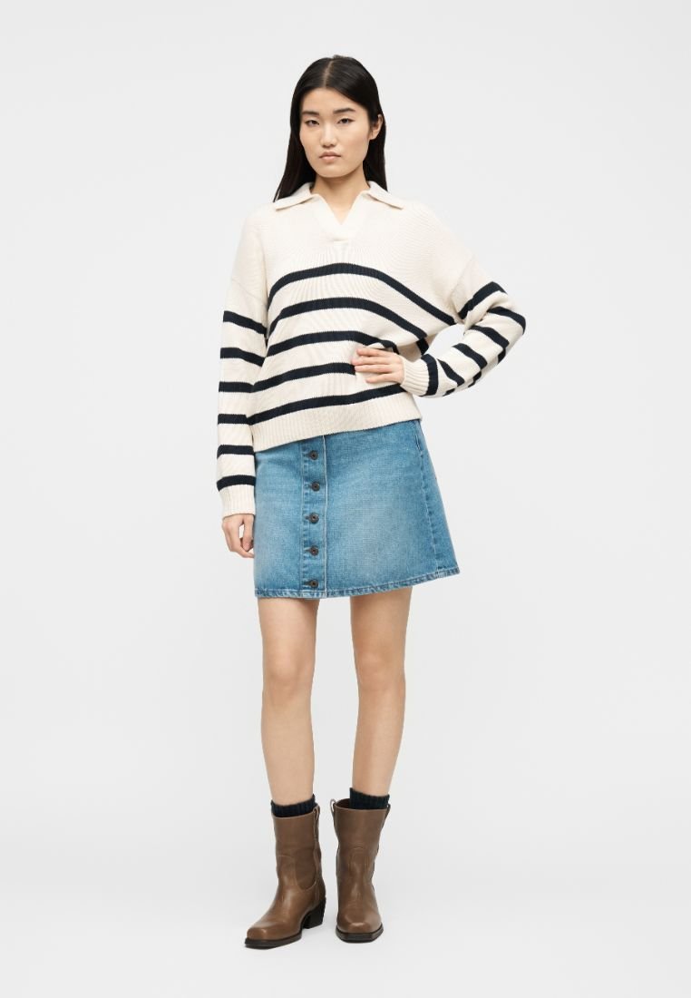 Jeune femme debout, main sur la hanche, portant un pull crème à rayures noires, une jupe en jean boutonnée, des chaussettes noires et des bottines marron.