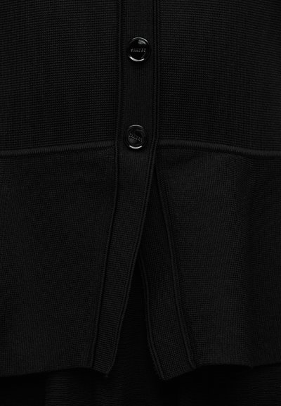 Κοντινό πλάνο μαύρου πλεκτού ζακέτα με δύο γυαλιστερές μαύρες κουμπότρυπες και εμφανή υφή πλέξης.