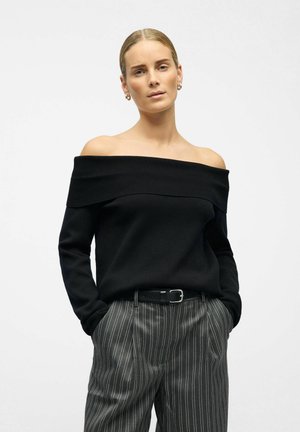 Pull noir en tricot décolleté, avec un col côtelé, associé à un pantalon gris à rayures et une ceinture noire. Texture lisse, design décontracté.