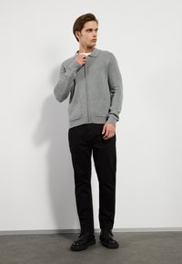 Pelēks zip-up kardigans ar apkakli, izgatavots no mīksta adījuma materiāla. Tam ir divas priekšējās kabatas un garas piedurknes, apvienots ar melnām biksēm un apaviem.