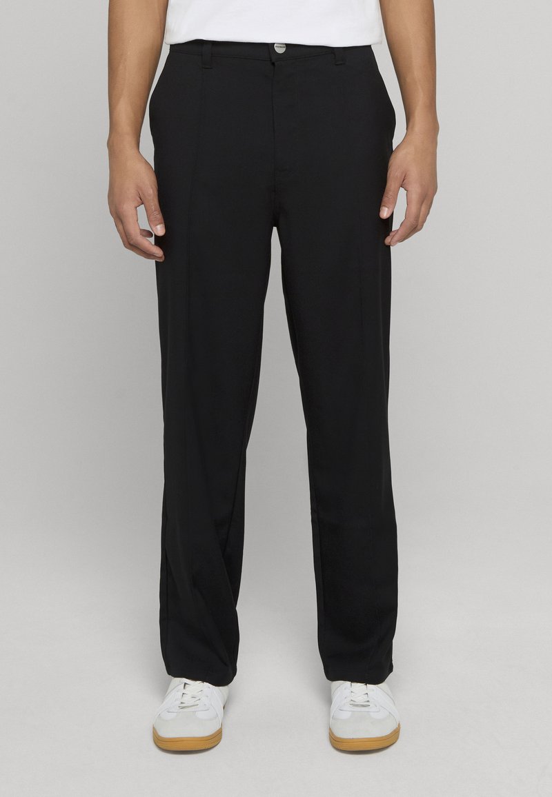 Černé kalhoty s úzkým střihem, hladkou látkou, předními záhyby a zapínáním na knoflíky, kombinované s bílými teniskami a jednoduchým tričkem.