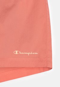 Pantalones cortos de color coral fabricados con un material ligero, con una textura suave, costuras laterales y un logo "Champion" de color crema en el dobladillo.