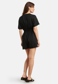 Black short-sleeve romper with elastic waist. Features a relaxed fit and lace trim at the hem. Paired with black block-heeled sandals.