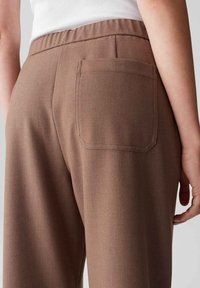 Femme portant un pantalon marron taille haute avec une poche arrière et une ceinture élastique, associé à un haut sans manches blanc.