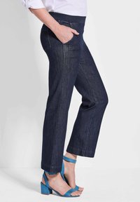 Jeans en denim bleu foncé avec une coupe décontractée, dotés d'une large ceinture et de poches avant, associés à des sandales à talons hauts teal.