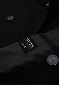 Czarna kurtka od wewnątrz z gładką teksturą, z nadrukowaną etykietą "ombre QUINTESSENCE rozmiar M" oraz srebrnym guzikiem.