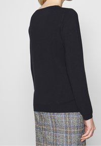 Schwarzer Pullover mit rundem Halsausschnitt und langen Ärmeln, aus einem weichen Material gefertigt. Trägt man über einem karierten Rock in Blau- und Brauntönen.