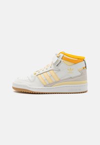 White high-top sneaker with yellow accents, leather and suede materials, three striped design on the side, rubber sole, and Velcro strap.