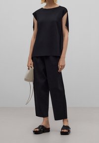 Nainen, jolla on päällään löysä musta hihaton paita ja leveät, lyhytlahkeiset housut, pitää käsissään beigeä ämpärilaukkua ja käyttää mustia avonaisia sandaaleja.