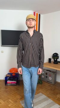 Camisa de botão às riscas em castanho-escuro e vermelho, combinada com jeans de perna reta azul-claro e um boné bege, situada num interior moderno.