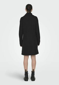 Robe en tricot noir avec un col montant, présentant un motif texturé, des manches longues et un ourlet qui tombe au milieu de la cuisse. Portée avec des bottines noires.