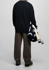 Pull en maille noire, pantalon ample marron, et chaussures foncées avec des lacets verts. Tient une veste universitaire bleu marine et crème avec des poignets rayés.