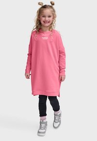 Rosa sweatshirtklänning med långa ärmar, har en logotyp på bröstet, kombinerad med svarta leggings och grå sneakers med vita detaljer.