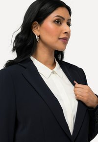 Blazer marine avec un design structuré, doté d'un col cranté et d'un tissu lisse. En dessous, une chemise blanche à col avec un détail décoratif.