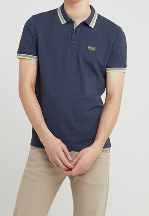 Man draagt een donkerblauw Hugo Boss poloshirt met witte en gele bies aan de kraag en mouwen, gecombineerd met beige broek.