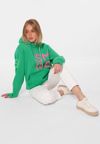 Felpa verde con il testo rosa "SMILE", cappuccio con cordini e tasca frontale. Indossata con pantaloni bianchi e scarpe chiare, seduto su uno sfondo neutro.