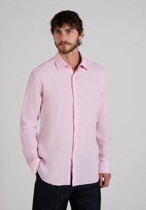 Hombre con barba vistiendo una camisa de manga larga color rosa claro abotonada y pantalones negros, de pie con las manos en los bolsillos contra un fondo liso.