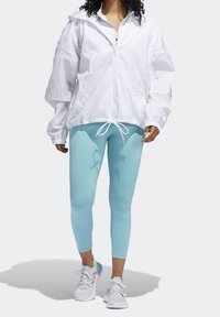 Giacca leggera bianca con zip e cappuccio, abbinata a leggings azzurri e scarpe da corsa multicolori. Caratterizzata da polsini elasticizzati e cordino.