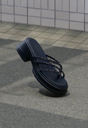 Schwarze Plateausandale mit drei dünnen Riemen und silbernen Schnallen, schwebt über Asphalt in der Nähe einer gefliesten Wand.