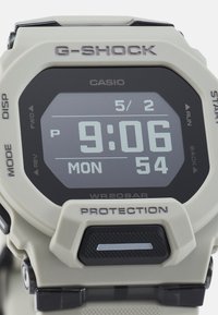 Montre numérique G-SHOCK de couleur beige clair avec un affichage noir. Présente un grand affichage numérique de l'heure, des boutons sur le côté et des accents texturés.