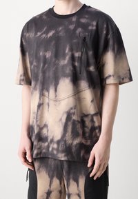 Camiseta de manga corta en un patrón de tie-dye negro y beige, hecha de tejido suave. Presenta un bolsillo en el pecho con cierre de cremallera y detalles cosidos.
