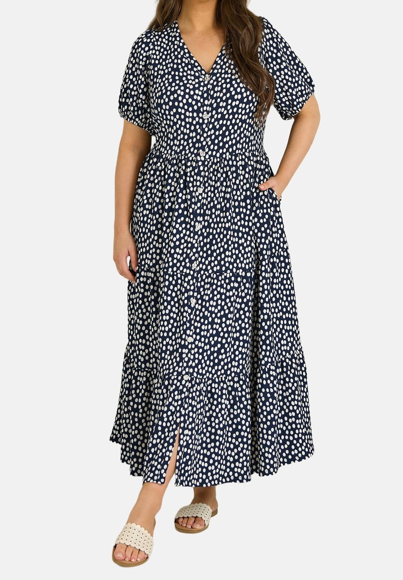 Femme portant une robe maxi bleu marine à pois blancs, manches courtes, boutons sur le devant, poche latérale et sandales blanches à enfiler.