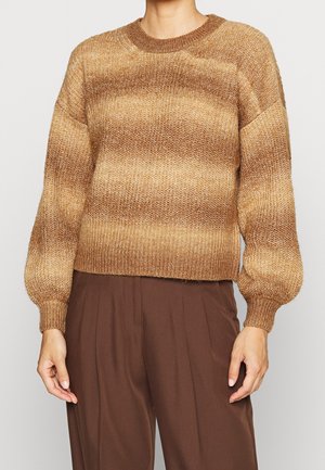 Personne portant un pull tricoté rayé marron et beige avec des manches bouffantes et un pantalon marron taille haute.