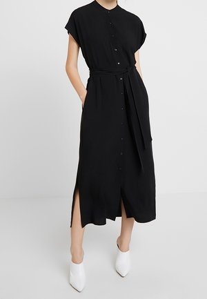 Žena stojící před jednobarevným pozadím, oblečená v černých midi šatech s knoflíkovým zapínáním vpředu, bočními rozparky a páskem v pase, na nohou má bílé kotníkové boty s špičatou špičkou.