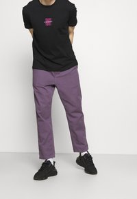 T-shirt en coton noir avec un logo graphique coloré, associé à un pantalon en coton violet et des baskets noires. Design simple, sans motifs visibles.