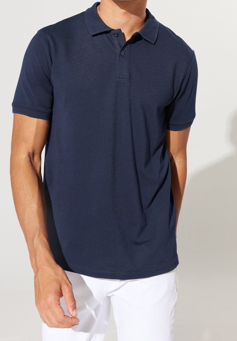 Homme portant un polo bleu marine à manches courtes et un pantalon blanc, debout devant un fond clair uni.