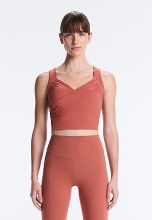 Nő rozsdavörös színű ujjatlan crop topban és hozzá illő, magasított derekú leggingsben, egyszínű világosszürke háttér előtt áll.