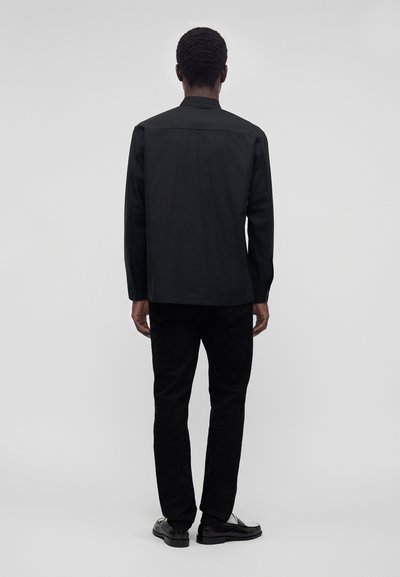 Homme debout, de dos, portant une chemise noire à manches longues, un pantalon noir et des chaussures noires devant un fond gris uni.