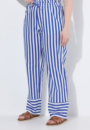 Persona con pantalones a rayas verticales azules y blancos con cintura con cordón y sandalias marrones, de pie sobre un fondo blanco.