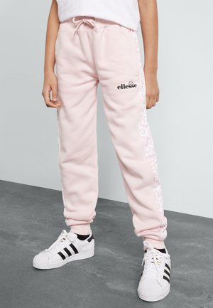 Personne portant un pantalon de survêtement Ellesse rose clair avec des bandes latérales à motifs et des baskets blanches Adidas avec des bandes noires sur un sol gris.