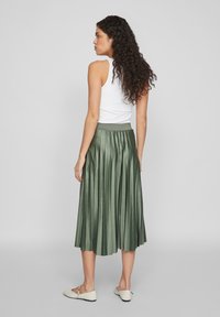 Gonna midi plissettata verde con una texture liscia e lucida. Presenta una vita elasticizzata e cade sotto i polpacci, abbinata a ballerine di colore chiaro.