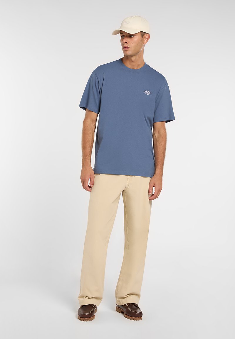 Maglietta di cotone azzurra con scollo rotondo e maniche corte. Pantaloni beige a gamba larga e scarpe marrone scuro. Completa il look un cappellino chiaro.