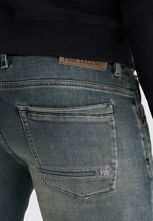 Wyblakłe niebieskie jeansy z denimu z przeszyciami na tylnych kieszeniach i brązową skórzaną metką PME Legend powyżej pasa, noszone z czarnym topem.