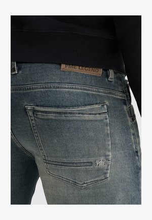 Verbleekte blauwe denim jeans met stiksel op de achterzak en een bruin leren PME Legend-label boven de tailleband, gedragen met een zwarte top.