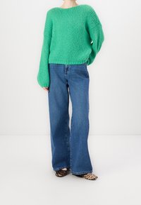 Pull vert en tricot avec des manches larges, associé à un jean bleu à jambes larges. Les chaussures présentent un motif léopard.