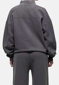 Felpa oversized grigio antracite con colletto alto, spalle cadute e polsini a coste. Tessuto morbido con una texture liscia.
