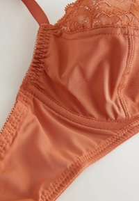 Krūšturis rudā oranžā krāsā ar gludu satīna materiālu, mežģīņu apdare augšā un smalkas šuves gar malām. Iekļautas regulējamas siksnas.