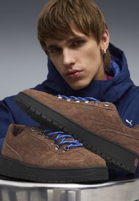 Brūni zamša apavi ar melniem zolemi, ar zilām auklām un metāliskiem aciņiem, teksturēta virsma un iegravēts logo. Parādīti no leņķa.