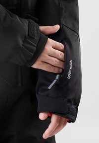 Detail einer schwarzen wasserdichten Jacke mit einem enganliegenden Bündchen mit Klettverschluss, das eine Tasche mit Reißverschluss und reflektierendem Akzent freilegt.