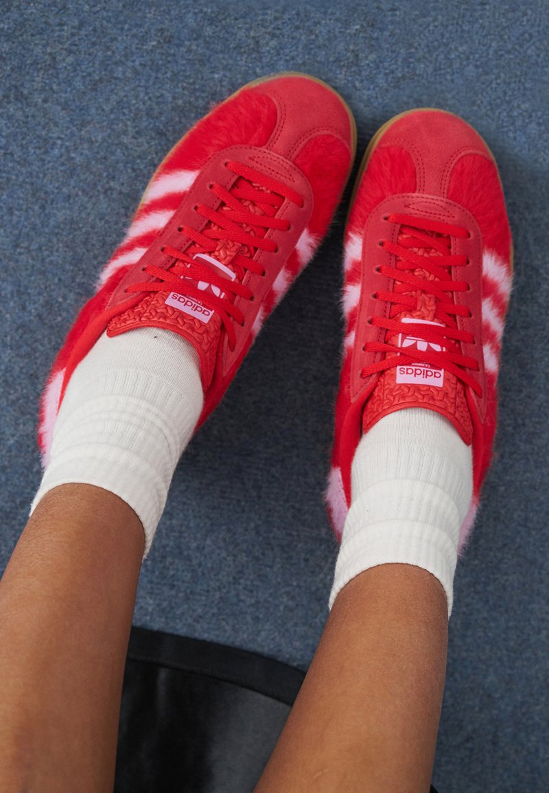 Pés calçados com ténis Adidas vermelhos brilhantes com riscas brancas e detalhes texturizados, combinados com meias brancas de cano médio numa carpete azul.