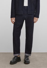 Tummansiniset farkkutakki ja housut, suoralla leikkauksella, napillinen sulkeminen ja käännetyt hihansuut. Yhdistetty mustiin nyörikenkiin.